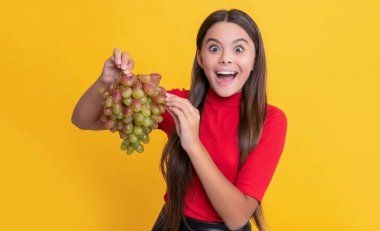 Afallamış genç kız gülümsüyor sarı arka planda bir demet üzüm tutuyor
