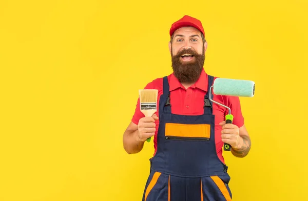 Erstaunt bärtiger Mann Hausmaler in Arbeitskleidung halten Farbroller und Pinsel auf gelbem Hintergrund — Stockfoto