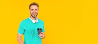 Mutlu, traşsız, kulaklıklı genç adam sarı arka planda kahve içiyor.