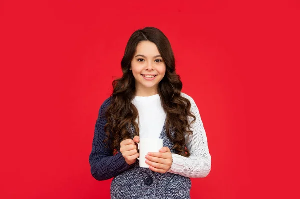 Criança sorridente com cabelo encaracolado em suéter de malha. menina adolescente beber chá quente no fundo vermelho. — Fotografia de Stock
