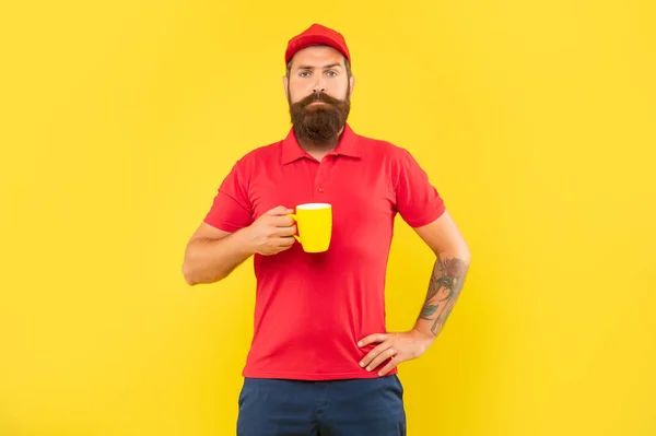 Grave ragazzo barbuto in casual berretto rosso e maglietta tenendo tazza sfondo giallo, caffè — Foto Stock