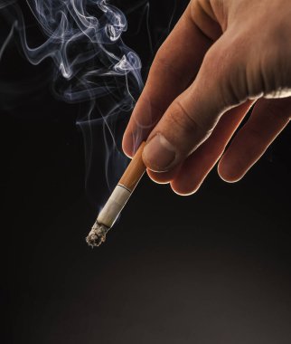 Male hand hold cigarette butt with smoke swirls dark background, copy space, nicotine addiction