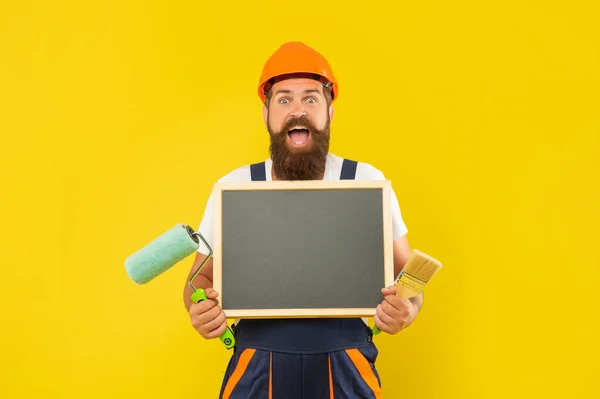 Užaslý muž v helmě a pracovní oblečení držet barvu váleček štětec a tabule s kopírovacím prostorem — Stock fotografie