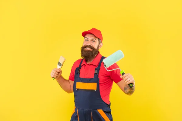 작업복을 입고 행복 한 턱수염을 가진 남자가 노란 배경에 페인트 롤러와 붓을 잡고 있다 — 스톡 사진