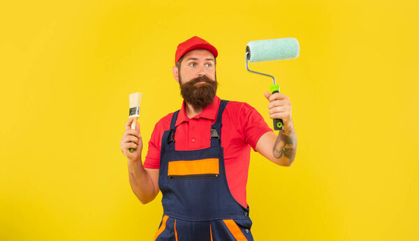 puzzled bearded man in work clothes hold paint roller and brush on yellow background