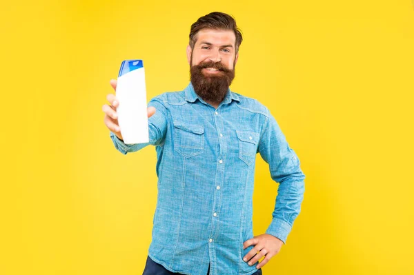 Brutal bearded man hold hair conditioner bottle on yellow background, presenting toiletries — Foto Stock