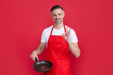 happy mature man chef in apron with frying pan on red background. ok