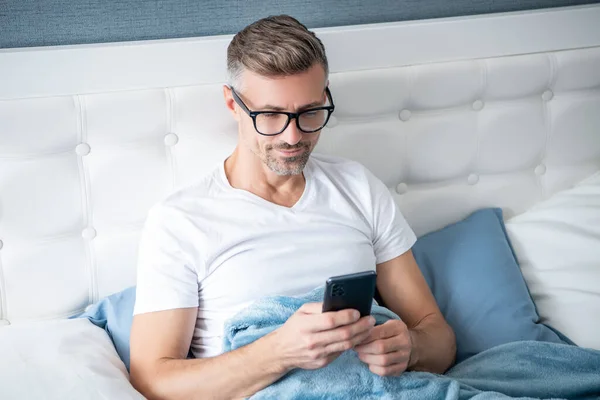 Grizzled man in bed chatting on phone wearing eyeglasses —  Fotos de Stock