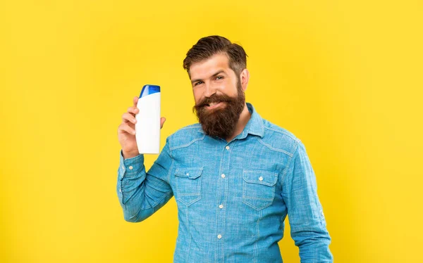 Cheerful brutal bearded man hold shampoo bottle on yellow background, presenting toiletries — Foto Stock