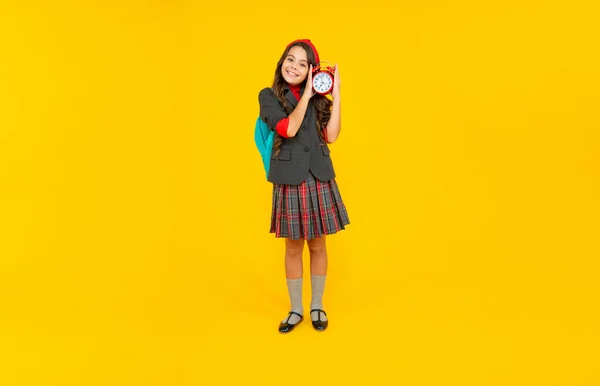 Chica adolescente feliz en uniforme con bolsa de la escuela mantenga el reloj despertador en el fondo amarillo, estudiante —  Fotos de Stock