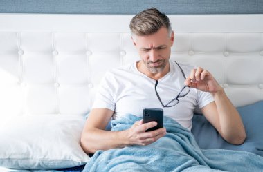 Yatakta konsantre olmuş bir adam, gözlüklü bir telefon okuyor.