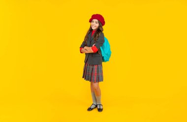 Sarı arka planda beresi ve sırt çantası olan okul üniformalı mutlu bir çocuk..