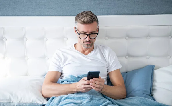 mature man in bed check email on phone wearing eyeglasses.