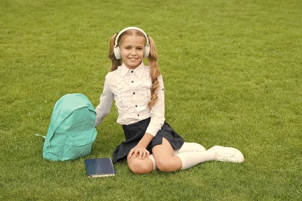 制服を着た幸せな子供は 緑の芝生の上に座ってヘッドフォンで音楽を聴く 学校の休日 — ストック写真