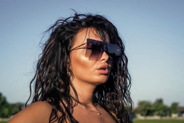 Seksi esmer kadın uzun kıvırcık saçlı şık güneş gözlüğü takıyor ve yaz havasının, modanın ve güzelliğin tadını çıkarıyor..