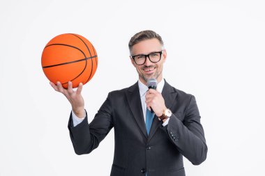 Takım elbiseli neşeli olgun adam basketbol topu ve mikrofonu beyaz arka planda izole etti..