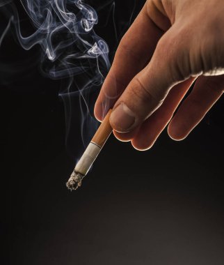 Male hand hold cigarette butt with smoke swirls dark background, copy space, nicotine addiction.