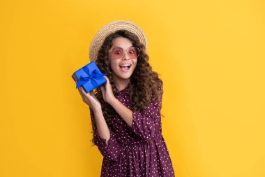 Kıvırcık saçlı mutlu çocuk sarı arka planda hediye kutusunu tutuyor..