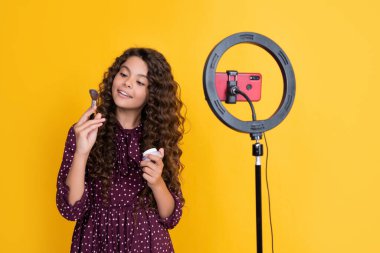 Uzun kıvırcık saçlı, gülümseyen çocuk blogcu blog tutuş pudrası fırçası videosu yapıyor.