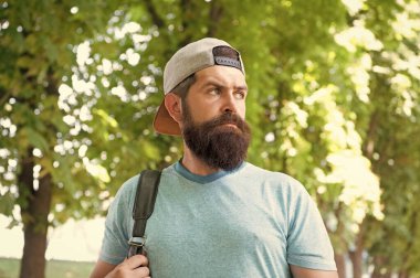 Sakallı ve bıyıklı, yaz tişörtlü ve şapkalı olgun sakallı bir adam ormanda yürüyüş yaparken sırt çantasını tutuyor..