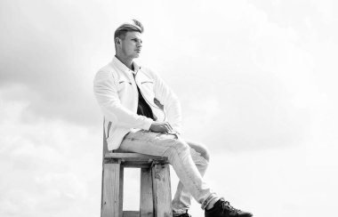 Hem gündelik hem de modaya uygun giyinmek. Yakışıklı adam açık havada rahat giyiniyor. Adam bulutlu gökyüzünde sandalyeye oturur. Günlük kıyafet dolabı. Moda trendleri. Moda tarzı. Erkek giyim mağazası. Günlük giysiler..