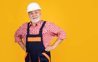 Sarı arka planda sert şapkalı mutlu kıdemli inşaatçı..