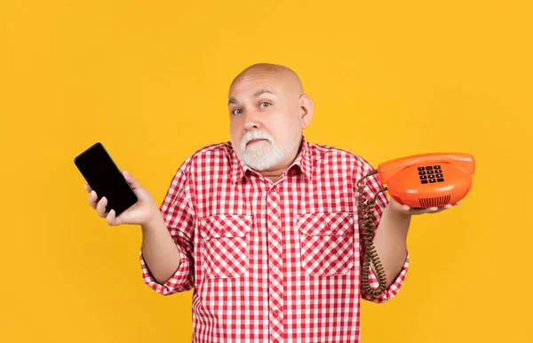 confused old man with retro telephone and modern smartphone on yellow background.