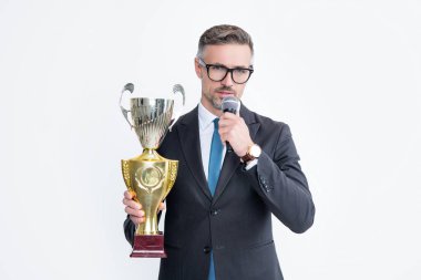 Takım elbiseli olgun bir adam şampiyon kupası ve beyaz arka planda izole edilmiş mikrofon tutuyor..