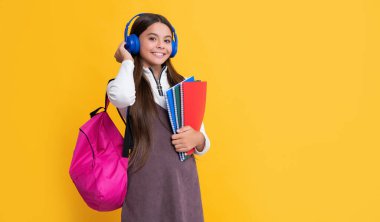 happy kid in headphones with school backpack with workbook on yellow background.