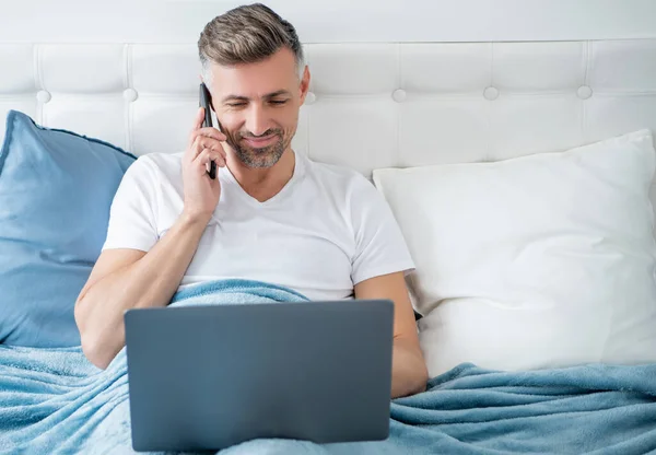 mature man speaking on phone and working on laptop in bed.