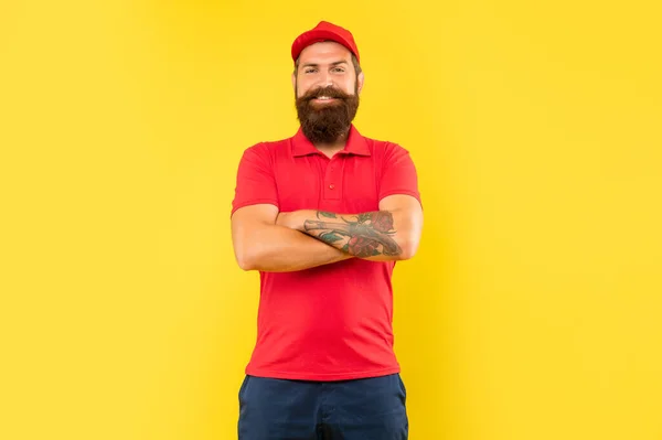 Happy confident guy in casual red cap and tshirt keeping tattooed arms crossed yellow background, confidence.