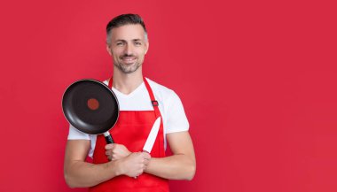 glad mature man chef in apron with frying pan and knife on red background.