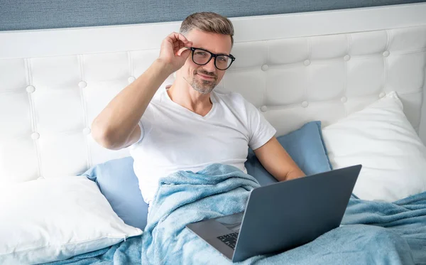 mature man in glasses check email on laptop in bed.