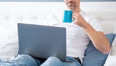 Neşeli olgun bir adam yatakta bilgisayarla kahve içiyor..