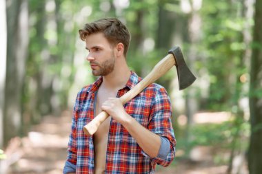 Keresteci gömlekli traşsız bir adam elinde baltayla ormanın arka planına bakıyor..