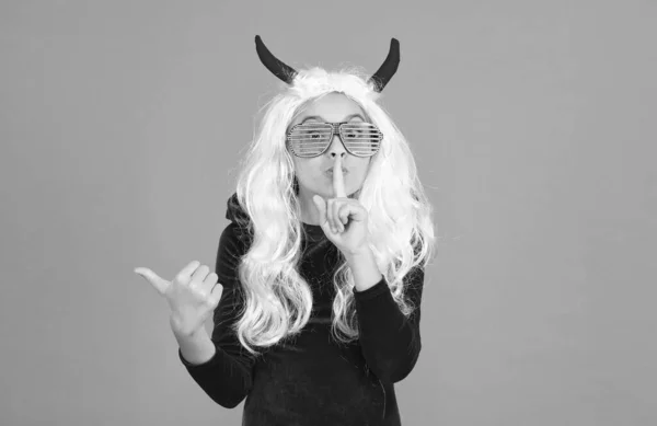 girl wear devil horns and party glasses having long white hair wig pointing finger and keep a secret with hush gesture, happy halloween secret gossip.