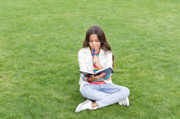 pondering teen girl making notes in notebook sitting on grass. taking notes. student make notes outdoor.