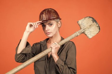 Koruyucu miğfer ve bikini giyen kadın işçiler turuncu arka planda kürek tutuyorlar..