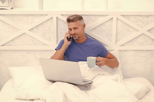 Happy man talk on mobile phone drinking coffee and using laptop in bed in morning, communication.
