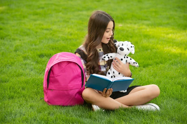 Teen girl reading book to dog toy sitting on grass, school. back to school. pupil at school time.