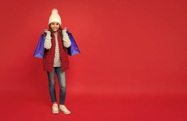 Kış şapkalı gülümseyen çocuk kırmızı arka plandaki alışveriş çantalarını tutuyor, fotokopi alanı ile kapatıyor..
