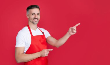 happy mature householding man in apron on red background. pointing finger.