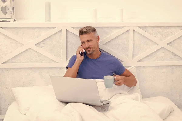 Mobile talking. Happy man talk on mobile phone in morning. Drinking coffee and using laptop in bed. Got phone call.