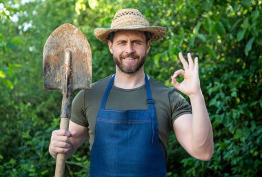Happy farmer man in farmers hat and apron holding garden spade and showing OK ring gesture natural outdoors.