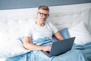 mature man virtual assistant in glasses working on laptop in bed.