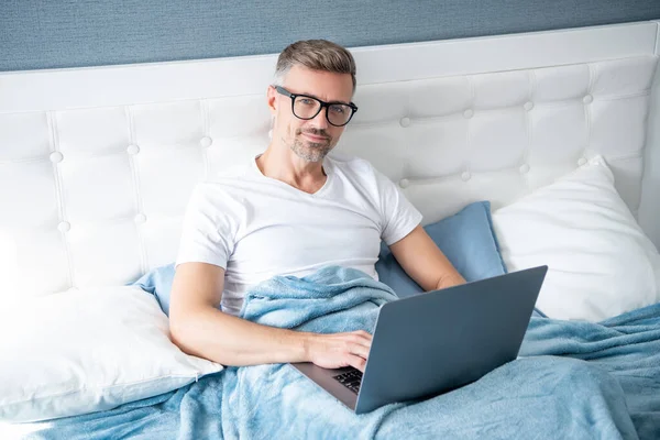 mature man virtual assistant in glasses working on laptop in bed.