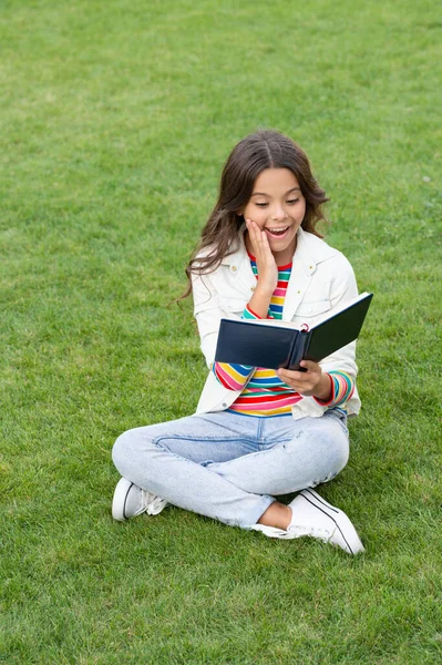 surprised teen child read book sitting on grass. reading book. reader girl with book outdoor.