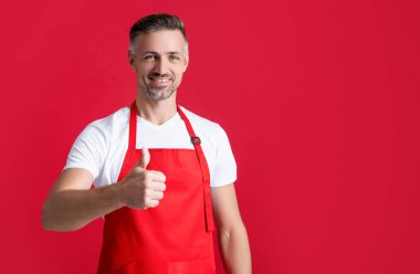 mature barista in apron on red background. thumb up.