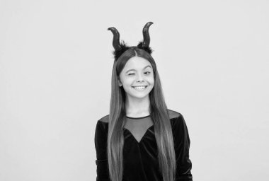 happy halloween. child in imp horns on yellow background. kid wear devil horns and winking. childhood autumn holiday. funny teen girl ready to celebrate party. costume party fun.
