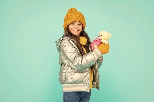 smiling teen girl hold toy bear. child wearing warm clothes on blue background. express positive emotion. winter fashion. love toy for valentines day. kid in puffer jacket and hat. childhood.